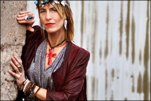 Woman with layered necklaces and a headband, posing against a textured wall.