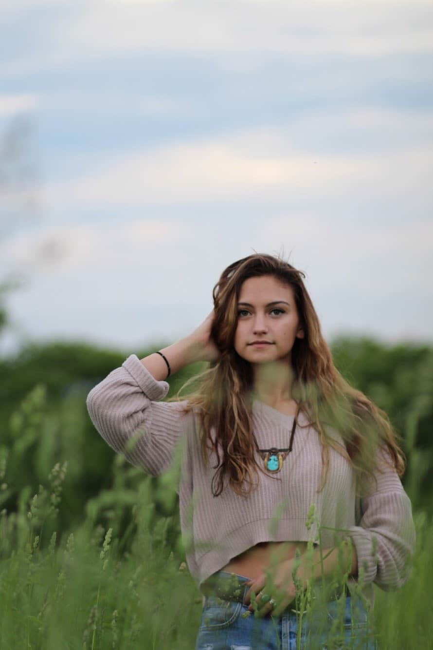 Young woman with long hair standing in a green field under a cloudy sky.