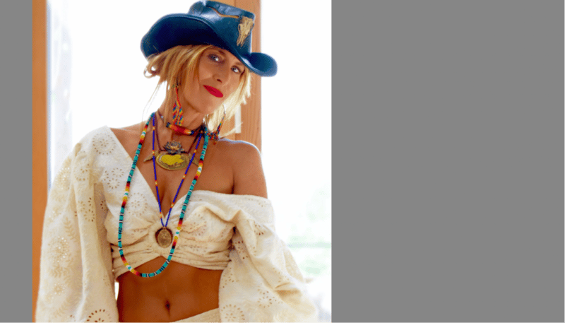 Woman in a blue hat and layered necklaces poses with a confident expression.
